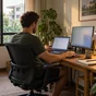 Ein Mann arbeitet an Laptop und zweitem Monitor in einem ruhigen Zimmer mit Blick auf tropische Pflanzen.