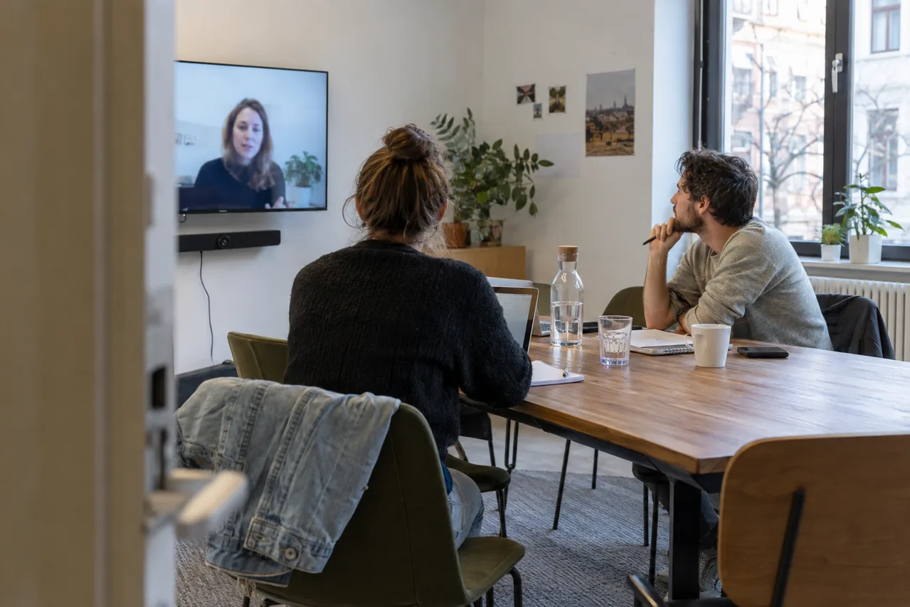 Zwei Mitarbeitende in einem Besprechungsraum mit einer weiteren Person per Videokonferenz.